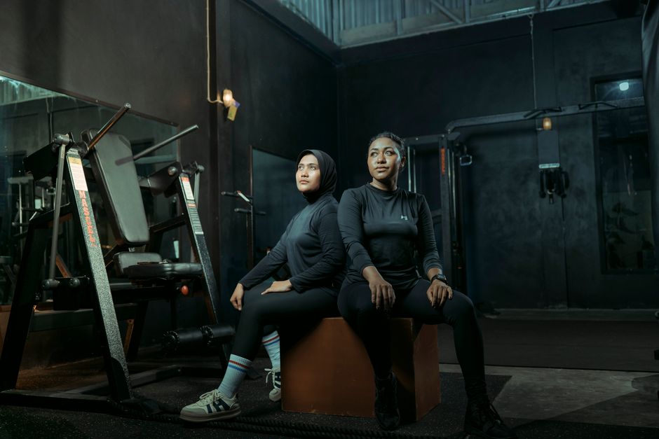 Two women in athletic wear sitting on a box with gym equipment in the background.